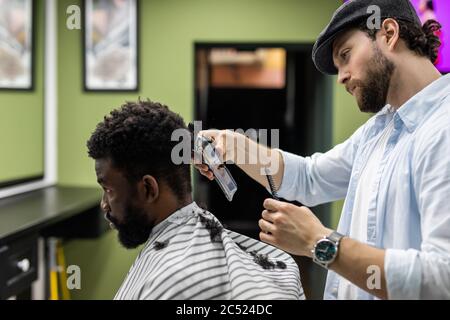 Closeup of process of trimming of hair in barber shop. Qualified barber keeping clipper in hands and correcting shape of hair to male client sitting o Stock Photo