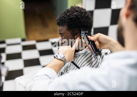 Closeup of process of trimming of hair in barber shop. Qualified barber keeping clipper in hands and correcting shape of hair to male client sitting o Stock Photo