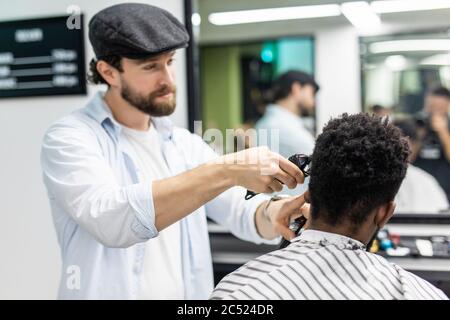 Happy young black man being trimmed with electric clipper machine in barbershop. Male beauty treatment concept. Smiling young African guy getting new Stock Photo