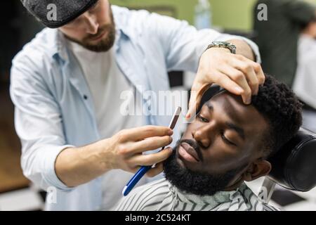 Side view of serious man with stylish modern haircut looking forward in barber shop. Hand of barber keeping straight razor and cutting trendy stripes Stock Photo