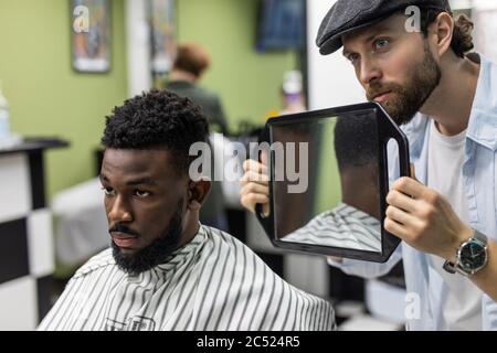 Portrait of handsome black man with comb in hair looking in the mirror at his new haircut. Barber hairdresser showing client his work. Male beauty tre Stock Photo