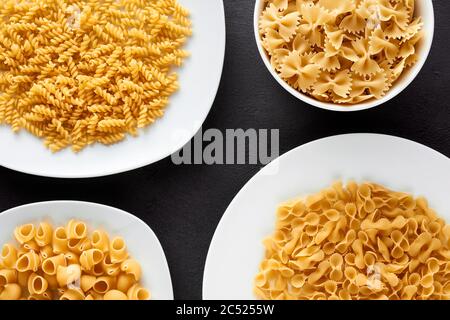 Variety of raw pasta in white plates and bowls on brown textured background. Flat lay view. Stock Photo