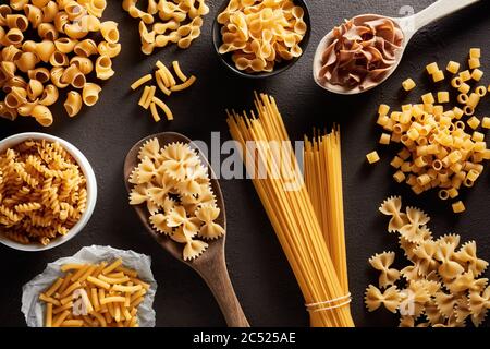 Heap of various raw pasta in different types and shapes on dark brown background. Overhead view. Stock Photo