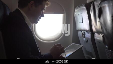 Young businessman traveling by plane and working. He sitting by the illuminator and typing message on tablet computer Stock Photo