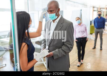 Woman with contactless fever measuring forehead at colleague with face mask in front of office because of Covid-19 Stock Photo