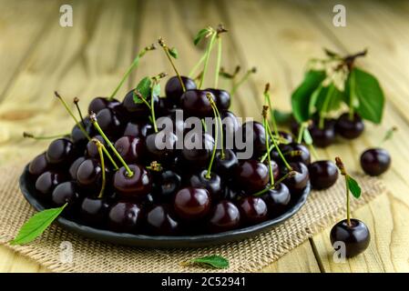 Fresh sour cherries on a black plate and green leaves on the wooden table. Fresh ripe sour cherries. Selective focus. Stock Photo
