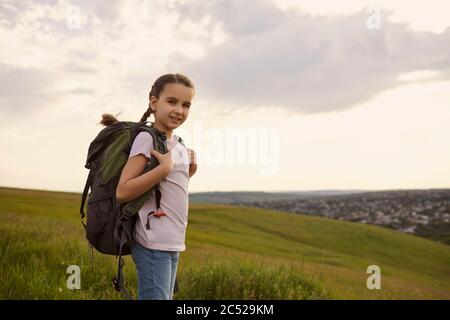 Adorable little girl with backpack standing on mountain top, copy space. Cute kid with rucksack hiking in countryside Stock Photo