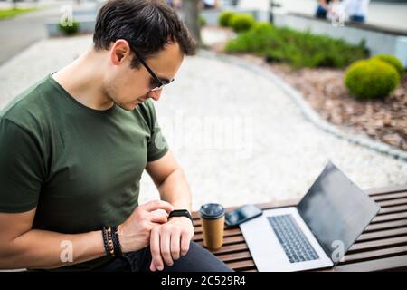 Stylish man sitting on bench with laptop and looking at watches. Stock Photo