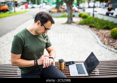Stylish man sitting on bench with laptop and looking at watches. Stock Photo
