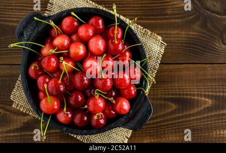Sweet red cherries in a black bowl on a brown wooden table with copy space for text. Top view. Stock Photo