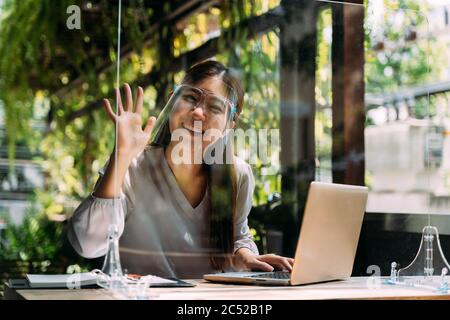 Smart young Asian woman smiling and waving hand while sitting at table with laptop and studying on terrace Stock Photo