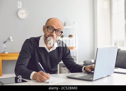 An elderly man with glasses is working he is studying he has a laptop he writes notes in a notebook while sitting at a table in his home office. Stock Photo