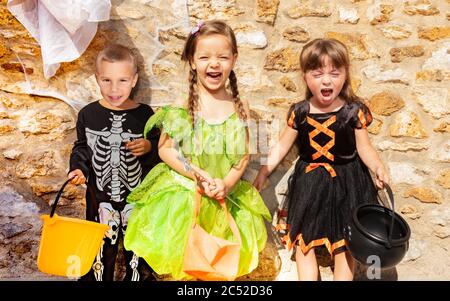 Group of little kids boy and two girls stand together in Halloween costumes, dresses over the stone wall with candy buckets Stock Photo