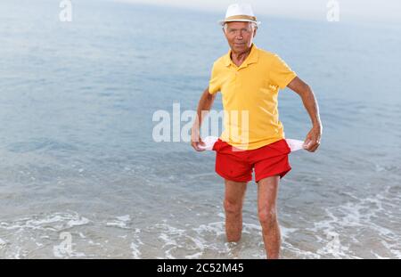 Sad mature man in headwear shows empty pockets near ocean Stock Photo