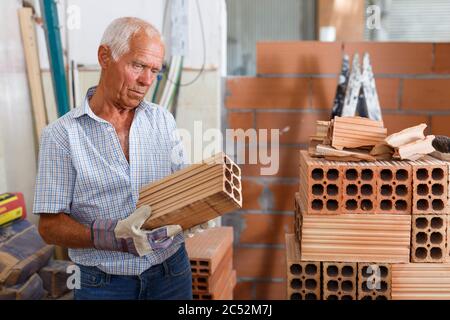Skillful older man inspecting brick for installing wall in building under construction Stock Photo