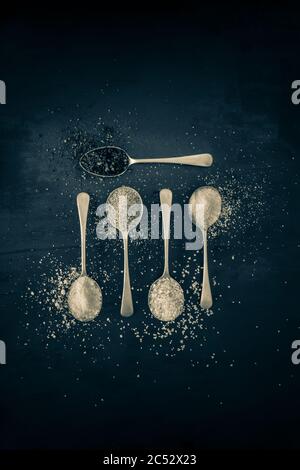 Antique silver spoons with various types of salts and spices laid out in patterns on a dark textured background Stock Photo