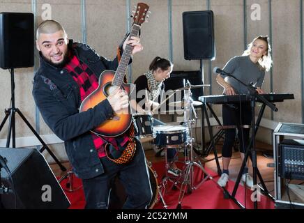 Bearded guy soloist playing the guitar and singing with two girls of his music band at the studio Stock Photo