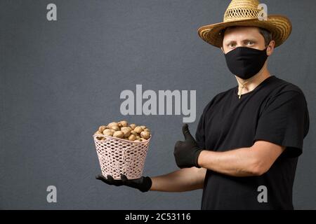 A farmer in a straw hat and a protective mask holds in his hand a wicker basket with walnuts. With the other hand shows a like. On a gray background. Stock Photo