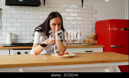 Young woman picks with fork in dish. Adult female sits thoughtfully in kitchen over plate of food. Stock Photo