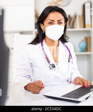 Focused latin female doctor in medical face mask attentively listening to patient complaints at clinic, writing prescriptio Stock Photo