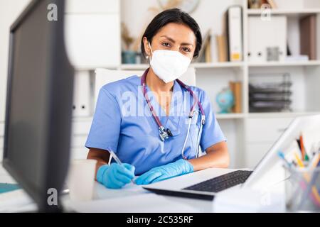 Focused latin female doctor in medical face mask attentively listening to patient complaints at clinic, writing prescriptio Stock Photo