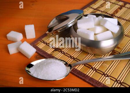 A bowl full of sugar cubes and a spoon full of refined white sugar on a wooden table. Stock Photo