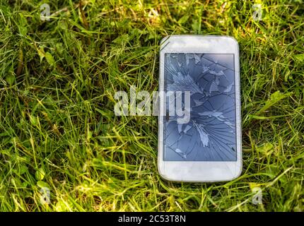 Broken mobile phone with cracked display on green grass background, outdoor, natural light. Stock Photo