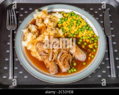 Overhead shot of dinner on a tray, with a knife and fork next to the plate of sausages, mashed potato, peas, sweetcorn, sliced onions and gravy. Stock Photo