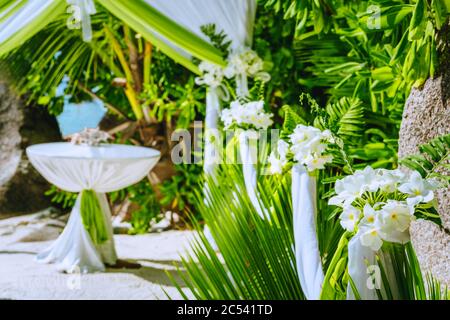 Decorated romantic wedding celebration location, table and chairs on tropical beach. Lush green foliage and white lowers. Stock Photo