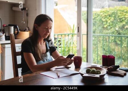 Young woman sitting on a wooden kitchen table with relaxed atmosphere checking her smartphone and writing notes; concept natural lifestyle, simplicity Stock Photo