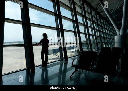 Female traveler looks out the window at the airport terminal. Young woman is waiting for boarding in the hall before departure. Stock Photo