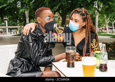 Two multi-racial, black and mixed-race friends on a bar terrace having a drink and hugging each other with face masks because of the coronavirus covid Stock Photo