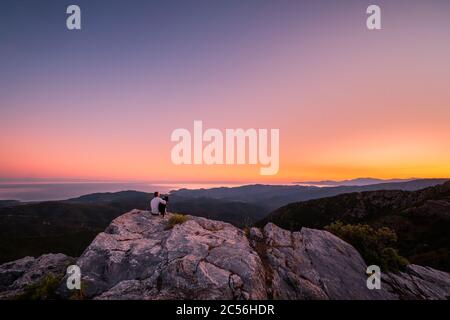 A man and his border collie dog enjoy the view of the coastline and the Mediterranean sea at sunrise from a rocky mountain outcrop at the Col de San C Stock Photo