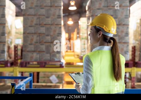 Side view of asian female warehouse worker with archboard flip chart look inventory with dustrubution warehouse in background. Reopening business ware Stock Photo
