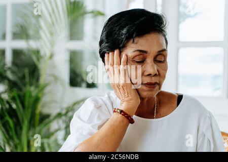 Stressed senior Asian female leaning on hand and pondering about problems at home. Elderly woman thinking about something. Stock Photo