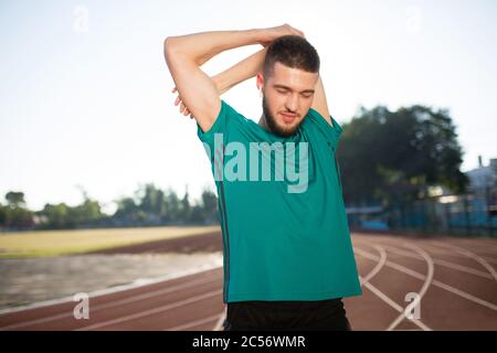 Young man in wireless earphones stretching arms on treadmill of stadium Stock Photo