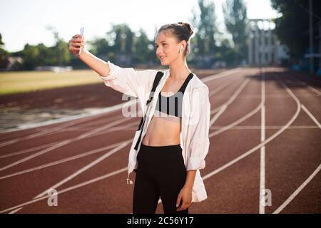 Beautiful smiling girl in earphones happily taking photo on frontal camera of cellphone while spending time on treadmill of stadium Stock Photo
