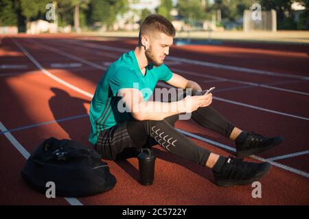 Young guy in wireless earphones thoughtfully using cellphone while spending time on running track of stadium Stock Photo