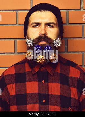 Bearded man with flowers or bows in long beard. Brutal caucasian serious unshaven hipster in black hat, checkered shirt on brick wall studio background. Party celebration concept Stock Photo
