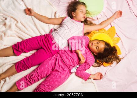 Kids in pink pajamas have fun. Schoolgirls having pajama party with funny pillows. Children with sleepy faces lie on light pink blanket background and hold hands up. Childhood and happiness concept. Stock Photo