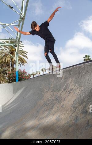 Young skateboarder performing tricks in a park, with street background and blue sky. Stock Photo