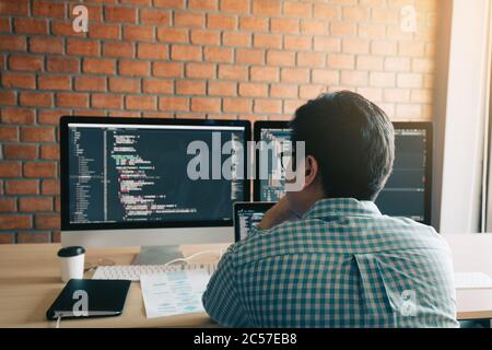 Rear view of contemporary website developer writing code for program and looking monitor screen with analyzing code. Stock Photo