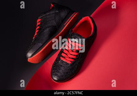 Fashionable snakeskin leather sneakers. Black sneakers with bright red bootlaces against a black and red background and long shadows. Stock Photo