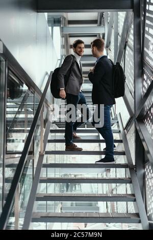 Two young businessmen going up, down the stairs while chatting in a modern building. Stock Photo