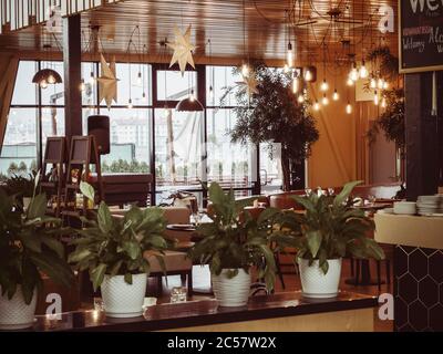 Russia, Sochi 14.03.2020. The interior of the cafe with plants standing on a stand in the foreground, many bright decorative lamps on a wooden ceiling Stock Photo