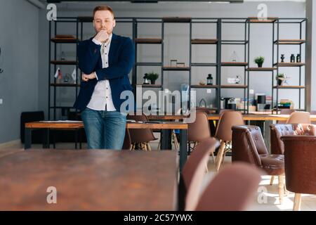 Thoughtful young man wearing fashion clothing is standing in modern office room near wooden desk. Stock Photo
