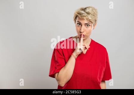 Portrait of young attractive female nurse making silence gesture on grey background with copy space advertising area Stock Photo