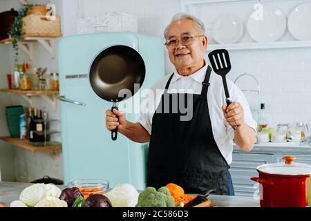 Happy senior Asian male in apron smiling for camera and showing frying pan and spatula while preparing lunch in kitchen at home Stock Photo