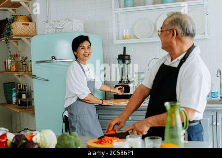 Senior Asian man and woman smiling and looking at each other. Elderly couple standing in cozy kitchen and cooking lunch together at home. Stock Photo