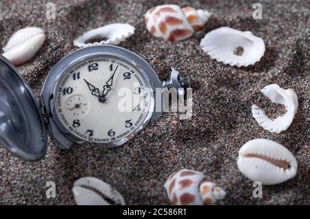 An old pocket watch lying in the sand surrounded by shells. Seashells. The clock fell in the sand. Time is running. Running time. Leaking time. Stock Photo